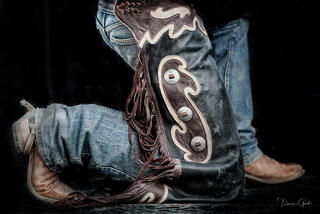 Close-up of ornately tooled leather cowboy chaps and worn boots against dark background, western fine art photography by Tamara Gooch.