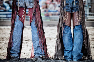 Color fine art photo of two cowboys standing side by side in ornate fringed leather rodeo chaps and jeans on a rodeo arena dirt floor.