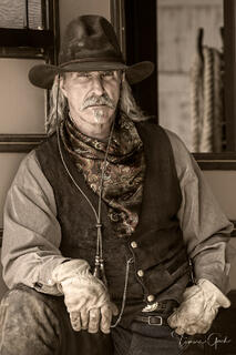 Weathered frontiersman in worn vest, paisley neckerchief and bolo tie, holding gloves, direct gaze, sepia western fine art portrait by Tamara Gooch.