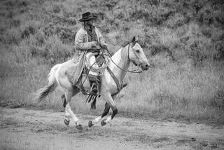 Black and white fine art photo of a cowboy in a duster coat riding a paint horse at a canter, rope coiled at saddle.