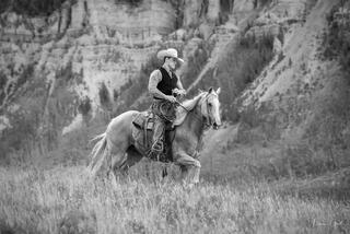 Black and white fine art photo of a cowboy on a pale horse riding through tall grass below dramatic layered sandstone canyon walls, American West.