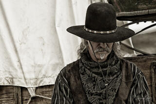 Weathered Cowboy in black hat with silver conchos, brocade vest and neckerchief, covered wagon background, sepia western fine art portrait by Tamara Gooch