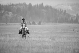 Black and white fine art photo of a lone cowboy riding toward camera across a vast misty mountain meadow, forested peaks behind.