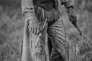Close-up of cowboy in chaps holding two syringes, worn leather gloves and fringed chaps, black and white western ranch fine art photography by Tamara Gooch.