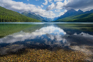 Cloud reflections on still waters of Bowman Lake, Glacier National Park, Montana, with colorful pebbles and forested mountains beyond.