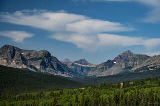 Wide valley view of the Continental Divide peaks rising above dense conifer forest, Glacier National Park, Montana, under a blue summer sky.