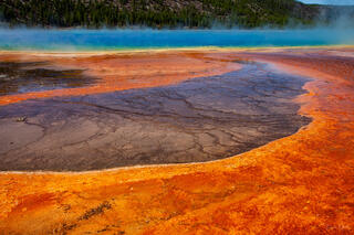 Color fine art photo of Grand Prismatic Spring's vivid orange and brown microbial mats at water's edge, turquoise center and steaming forest beyond, Yellowstone