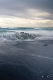 Glacier ice chunk surrounded by silky long exposure tide on Iceland's Diamond Beach black sand, stormy sky, teal water, fine art photography by Tamara Gooch.