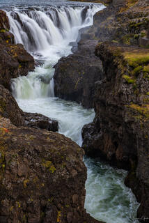 Waterfall cascading over basalt rim into churning turquoise gorge through volcanic canyon, Iceland.
