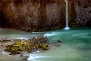 Long exposure Iceland fine art photograph of a thin waterfall falling into a turquoise river with mossy rocks.