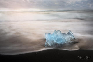 Single luminous blue glacier ice chunk on Iceland's Diamond Beach black sand, long exposure silky tide, soft warm sky, fine art photography by Tamara Gooch.