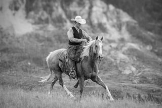 Black and white fine art photo of a cowboy in a vest and white hat riding a palomino at a trot, eroded badlands formations soft behind him.