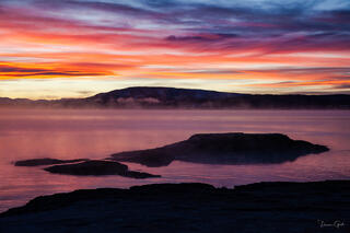 Fiery pink and orange sunrise sky reflected in the still waters of Yellowstone Lake, mist rising from thermal features along the silhouetted shoreline.
