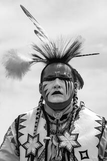 Black and white fine art portrait of a Native American pow wow dancer in full face paint, roach headdress, beaded regalia, against open sky.