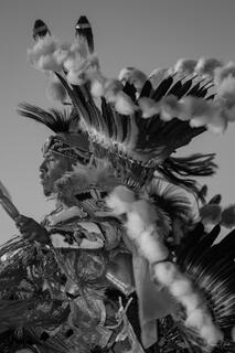 Black and white fine art photo of a Native American pow wow dancer in elaborate feathered war bonnet and regalia, staff in hand, against open sky.