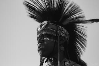 Black and white fine art portrait of a Native American pow wow dancer in profile, dramatic porcupine roach headdress rising against pale sky.