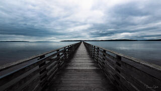 Long wooden pier extending to vanishing point over still grey Puget Sound, dramatic sky, distant tree-lined shores, fine art photography by Tamara Gooch.