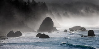 Oregon coast sea stacks rising through sea mist, dark evergreen forest behind,  blue-grey waves, shafts of light, panoramic fine art photography by Tamara Gooch