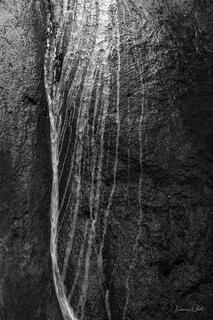 Black and white close-up photograph of water flowing and trickling down textured dark rock surface, abstract fine art nature photography print