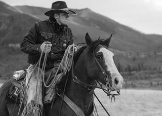 Black and white fine art portrait of a young cowboy coiling a rope on a paint horse, Wyoming mountains soft behind, focused expression.