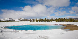 Color panoramic fine art photo of a vivid turquoise thermal pool surrounded by deep snow, conifers and blue sky behind, Yellowstone National Park.
