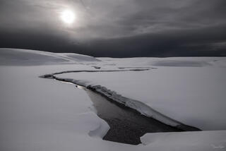 Dark winding creek through snow-covered Hayden Valley under dramatic storm sky, Yellowstone National Park, Wyoming.