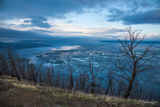 Aerial view of partially frozen Yellowstone Lake during spring ice melt, bare burned trees foreground, dramatic blue storm sky, landscape fine art photography 