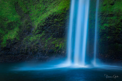 A lovely fine art limited edition print of a waterfall cascading into a pool of water. The dark rock wall behind covered with brilliant green foliage.