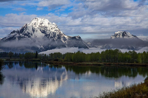 Color fine art photo of snow-capped Mount Moran reflected in still waters of Oxbow Bend, low clouds wrapping the range, spring cottonwoods, Grand Teton National
