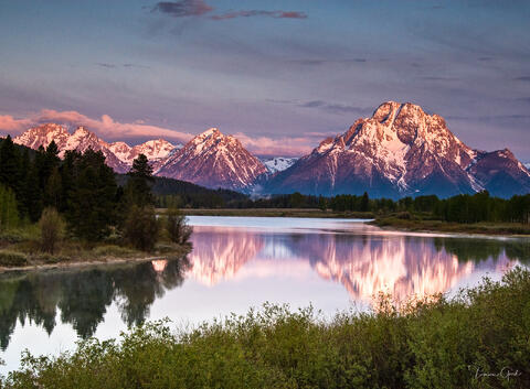 A wide fine art landscape of snow-capped mountains reflecting in a still river during a pink sunrise at Oxbow Bend.