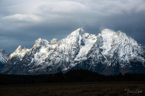 Mount Moran snow-covered peaks under dramatic storm clouds with shafts of light breaking through, dark conifer forest below, Grand Teton National Park landscape