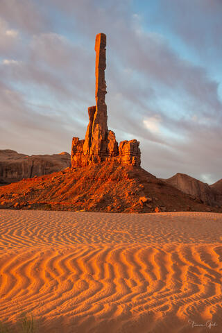 Totem Pole sandstone spire rising above glowing orange sand dunes at sunrise, Monument Valley Navajo Tribal Park.