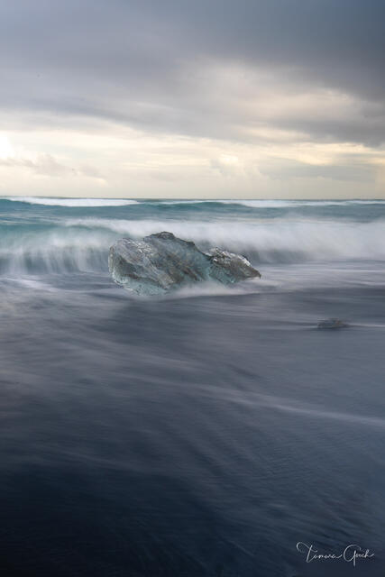 Glacier ice chunk surrounded by silky long exposure tide on Iceland's Diamond Beach black sand, stormy sky, teal water, fine art photography by Tamara Gooch.
