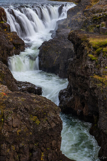 Waterfall cascading over basalt rim into churning turquoise gorge through volcanic canyon, Iceland.