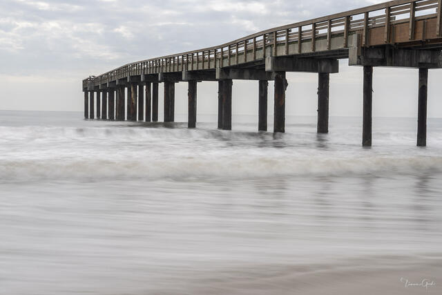 Florida pier extending diagonally across frame, long exposure silky surf around pilings, overcast sky, warm weathered wood, fine art photography by Tamara Gooch