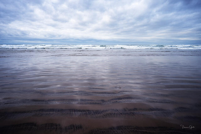 Wide Oregon beach at low tide, rippled wet sand reflecting moody blue-grey sky, distant waves on horizon, fine art photography by Tamara Gooch.
