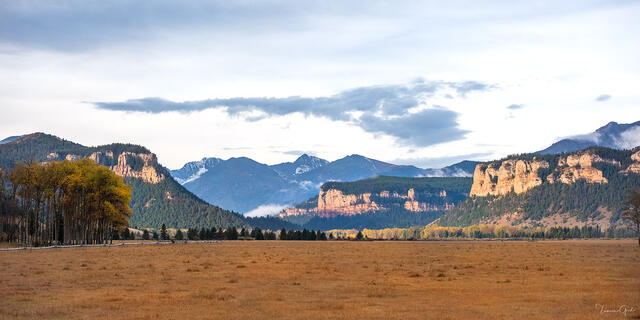 Golden autumn meadow stretching toward sunlit sandstone cliffs and mountain ridgelines under dramatic clouds, Sunlight Basin, Wyoming.