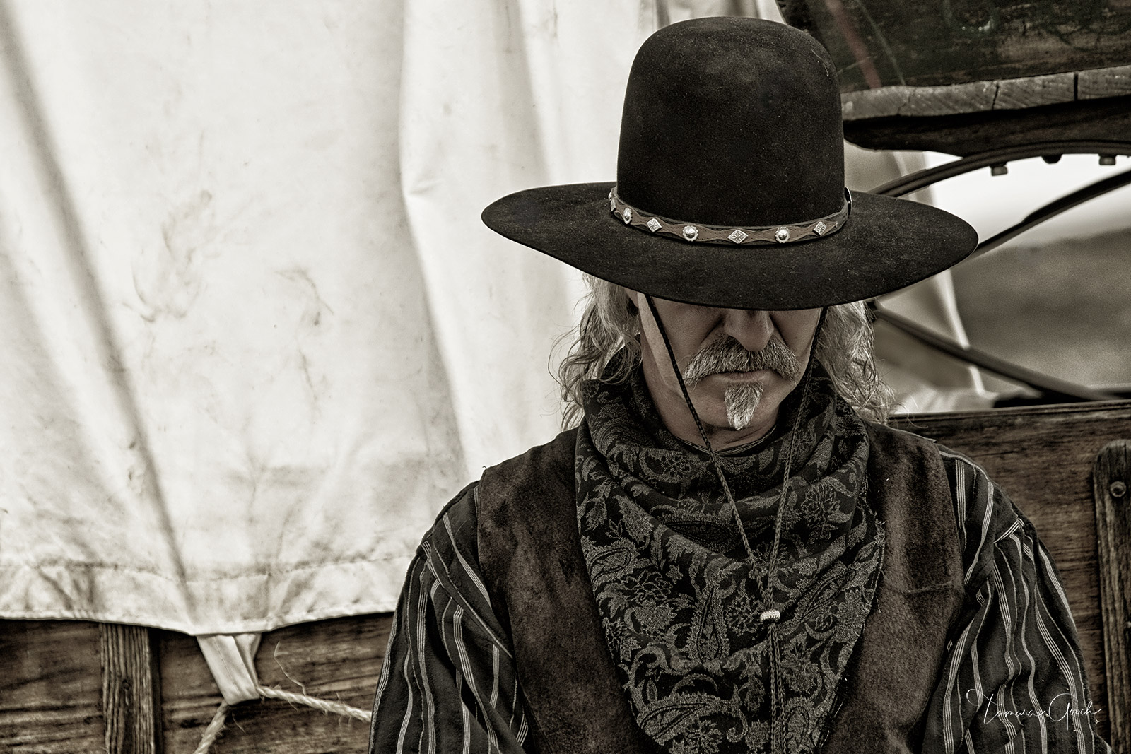 A rugged cowboy standing next to a covered wagon. Muted color. Limited edition print of the Western lifestyle.
